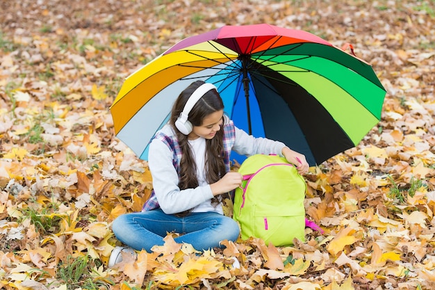 Bambino felice ascolta musica in cuffia mentre è seduto nella foresta autunnale tra foglie cadute con ombrello colorato e zaino scuola, autunno.