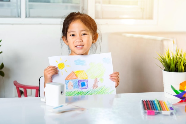 Bambino felice bambina sollevare paesaggio disegno colorato la mia casa sogno su carta per la telecamera asiatico carino bambino in età prescolare sorridente mostrando disegnare casa di campagna immagine a casa per imparare le arti compiti a casa