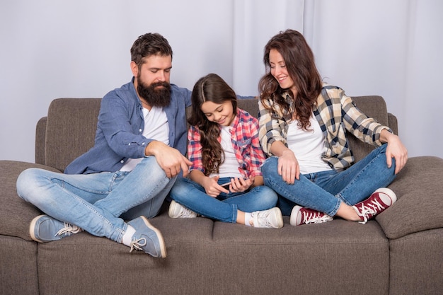 Bambino felice che gioca a giochi mobili su smartphone seduto accanto a padre e madre sul divano di famiglia