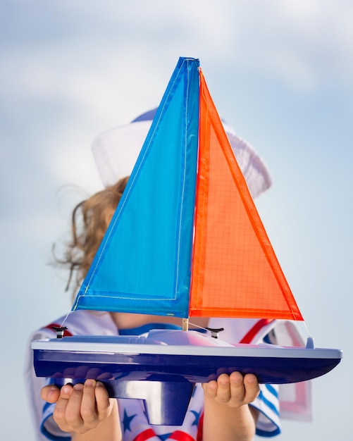 Bambino felice che gioca con la barca a vela del giocattolo contro il fondo del cielo blu