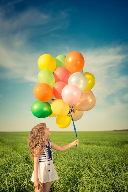 Bambino felice che gioca con palloncini colorati giocattolo all'aperto. Bambino sorridente che si diverte nel campo verde della primavera su sfondo blu del cielo. Concetto di libertà