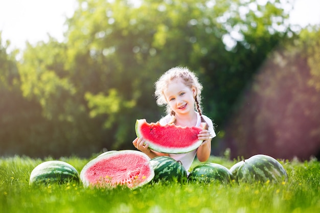 Bambino felice che mangia anguria. Il bambino mangia frutta all'aperto. Bambina che gioca in giardino morde una fetta di anguria.