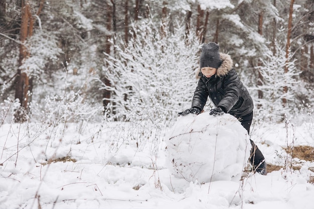 Bambino felice che rotola grande palla di neve per il pupazzo di neve nella foresta innevata dell'inverno. Ragazzo adolescente che gioca e si diverte a camminare in una giornata gelida. Attività invernale all'aperto.