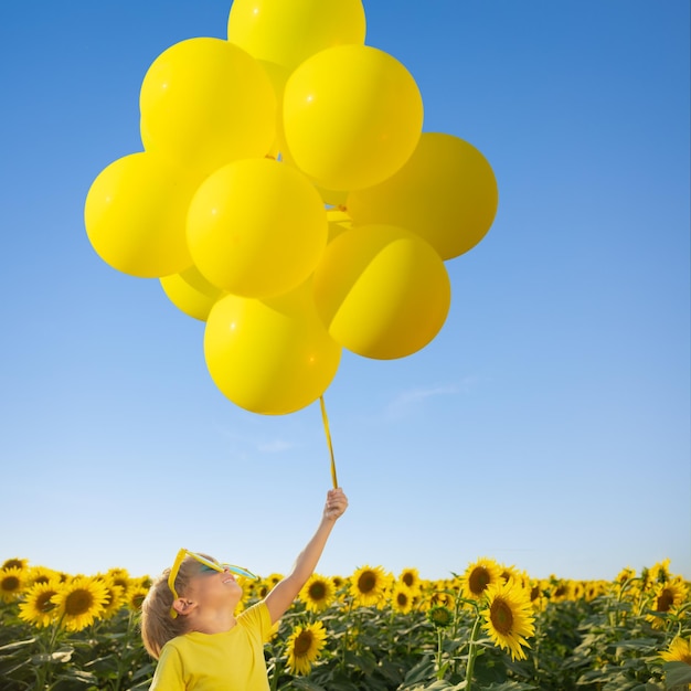 Bambino felice che si diverte nel campo primaverile dei girasoli. Ritratto all'aperto di bambino con palloncini colorati su sfondo blu cielo