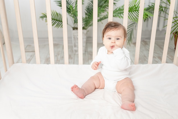 Bambino felice che si siede nella culla in una tuta bianca e succhia il dito