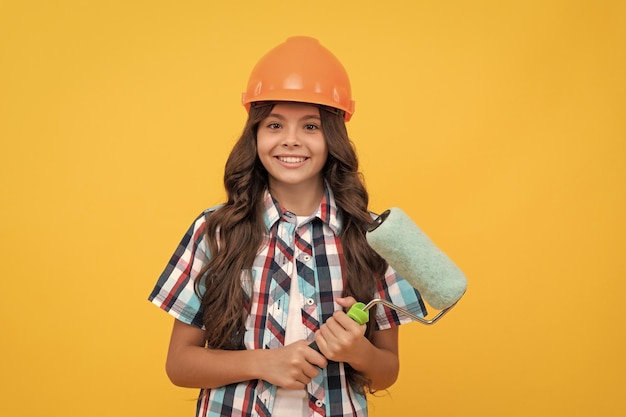 Bambino felice con i capelli ricci nella pittura del rullo di vernice della tenuta del casco della costruzione