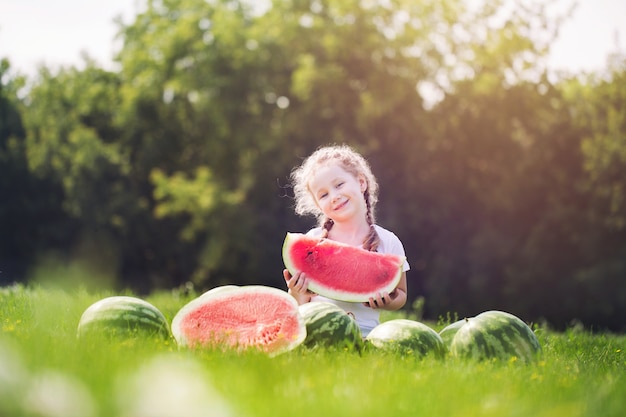 Bambino felice con la grande fetta rossa di anguria che si siede sull'erba verde nel parco estivo. Concetto di mangiare sano.