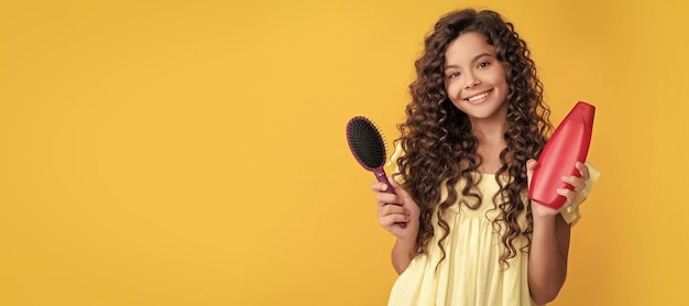 Bambino felice con lunghi capelli ricci, bottiglia di shampoo e spazzola per capelli. Cura dei capelli della ragazza del bambino orizzontale