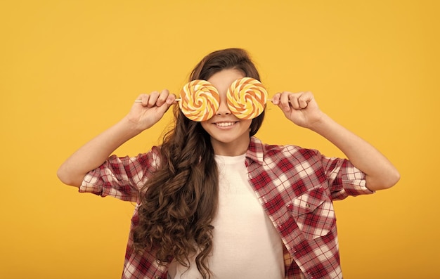 Bambino felice con lunghi capelli ricci in camicia tenere caramelle caramello lecca-lecca su sfondo giallo squisito