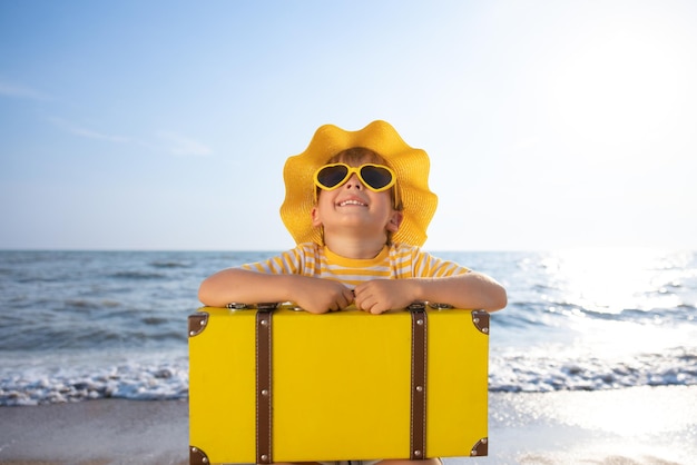 Bambino felice godersi il sole sullo sfondo del cielo blu Bambino con la valigia divertirsi sulla spiaggia Vacanze estive e concetto di vacanza