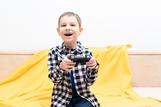 Bambino felice in camicia a quadri seduto sul divano con il joystick nero nelle sue mani giocando al videogioco Giocare ai videogiochi a casa concetto