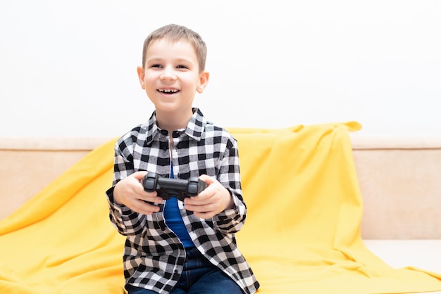 Bambino felice in camicia a quadri seduto sul divano con il joystick nero nelle sue mani giocando al videogioco Giocare ai videogiochi a casa concetto