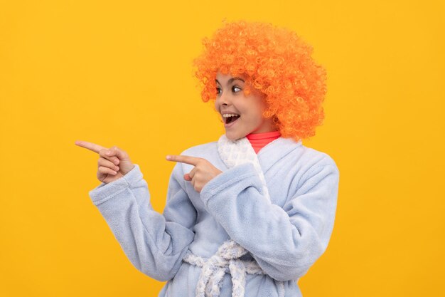 Bambino felice in parrucca arancione fantasia capelli indossare accappatoio da casa puntare il dito sullo spazio della copia, pubblicità.
