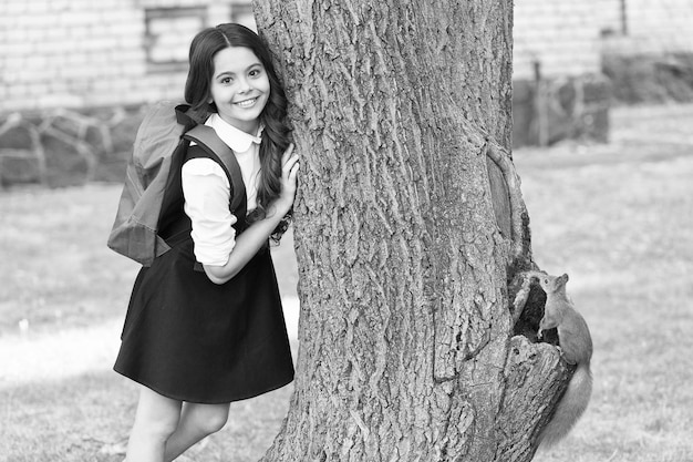 Bambino felice in uniforme con lo zaino della scuola ricerca scoiattolo che si arrampica sull'albero nella zoologia del parco