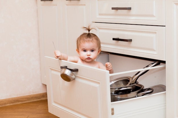 Bambino felice seduto nel cassetto della cucina con pentole e ridendo. Ritratto di un bambino in una cucina bianca.