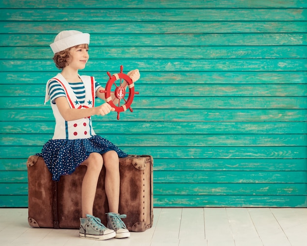 bambino felice seduto su una vecchia valigia vestito come un marinaio che guarda avanti su sfondo di legno blu