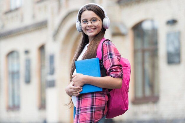 Bambino felice tenere il libro di scuola che ascolta il podcast audio in cuffie moderne all'aperto libreria musicale