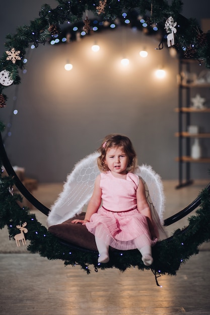 Bambino gioioso e felice che si siede e posa sulla corona di abete decorata con ghirlanda leggera. Adorabile ragazza che indossa in abito rosa carino e con ali d'angelo dietro. Kid sorridendo e guardando la fotocamera.