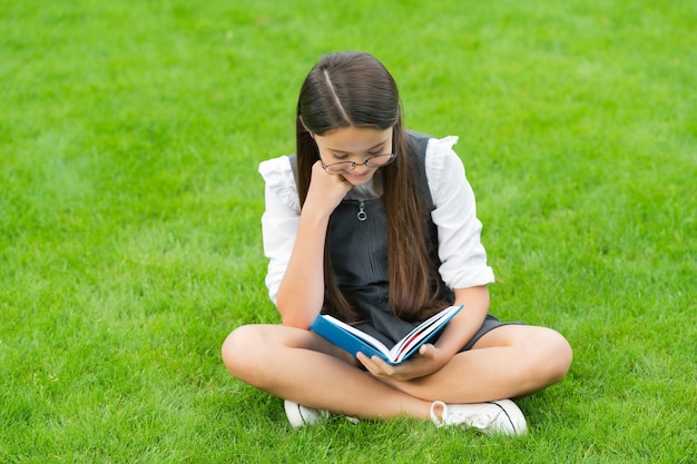 Bambino impegnato in occhiali che legge un libro seduto sull'erba verde