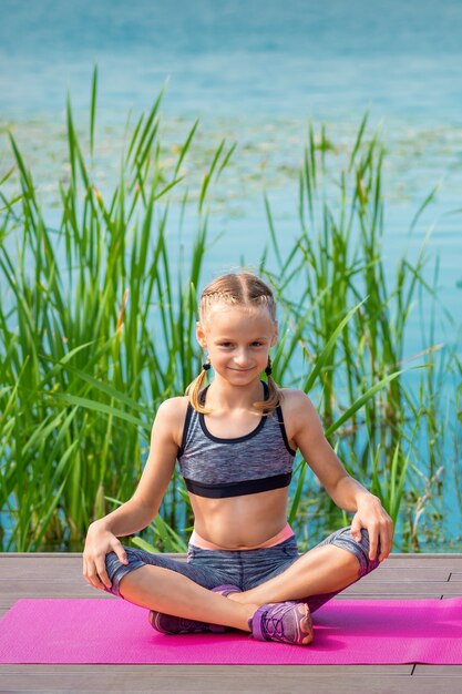 Bambino in abiti sportivi che si siede sulla stuoia sul molo vicino all'acqua all'aperto