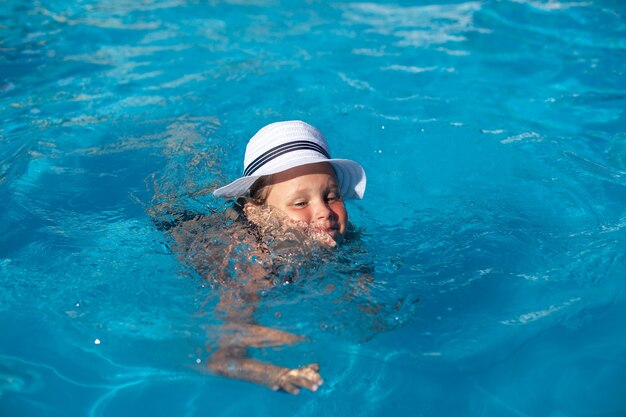 Bambino in cappello di paglia bianco felice ragazza affascinante sta imparando a nuotare in piscina e godendosi il riposo in estate calda...