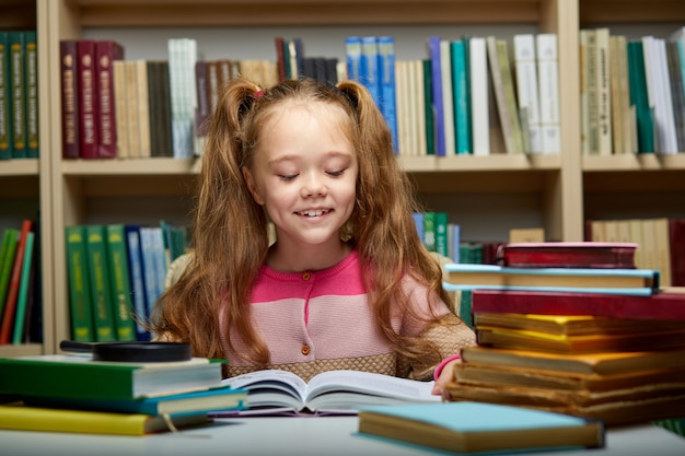 bambino in età prescolare bambina che legge un libro in biblioteca, piccola ragazza caucasica si siede con i libri vicino a una libreria