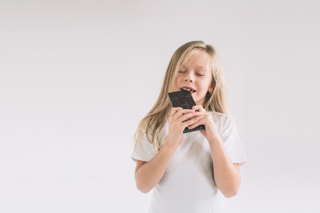 Bambino in giovane età che mangia una barra di cioccolato. Ragazza di Blondy isolata su bianco.