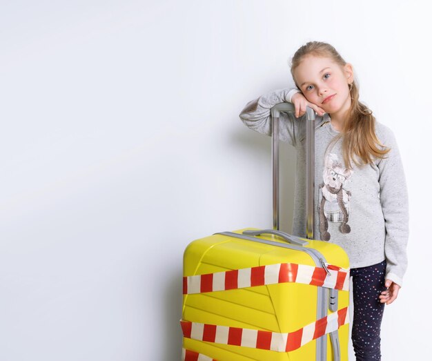 Bambino in maglione grigio in posa isolata su bianco Si appoggia su una valigia gialla legata da nastro di avvertimento bianco e rosso Coronavirus