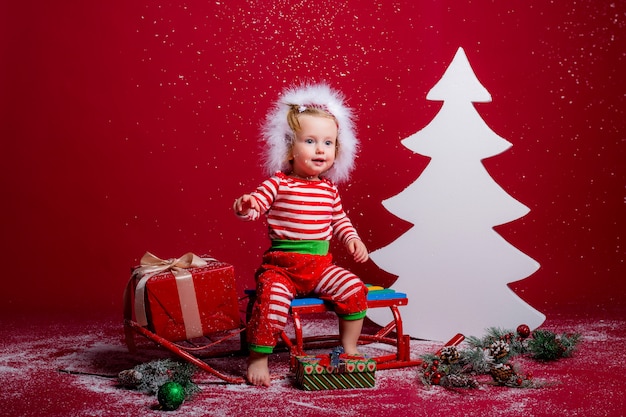 bambino in pigiama di Natale e cappello santa cattura neve seduto su una slitta con confezione regalo e grande albero di natale bianco su sfondo rosso