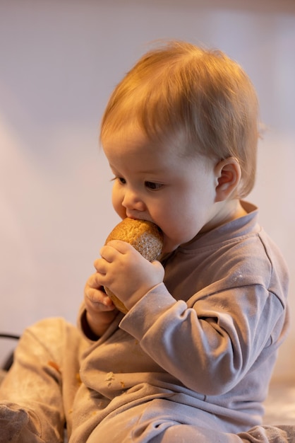 Bambino in tuta beige seduto sul tavolo nella cucina di casa e che mangia pane