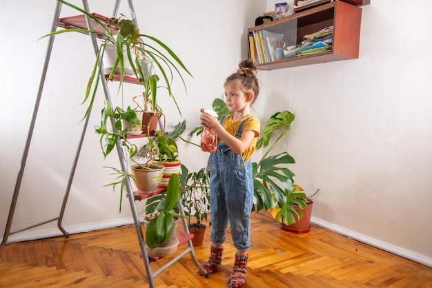 Bambino in tuta di jeans che schizzaxApiante d'appartamentoxAnella serra La bambina si prende cura delle piante verdi nella stanza Giardinaggio domestico amore per le piante e cura