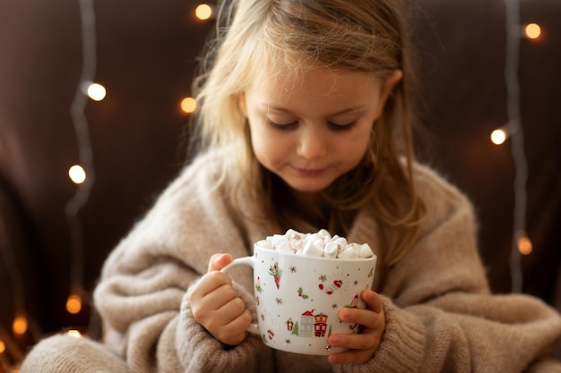 Bambino in un accogliente maglione lavorato a maglia tenere una tazza di cioccolata calda e luci ghirlanda di marshmallows