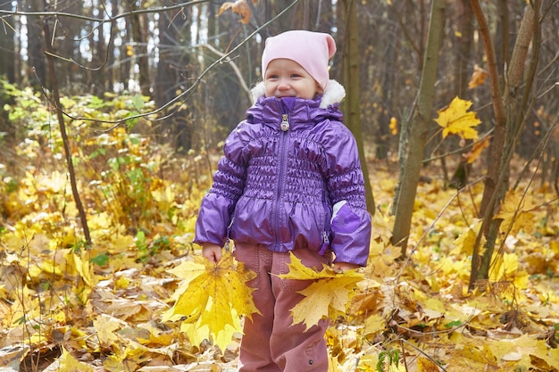 Bambino in una giacca viola con un cappello rosa con foglie d'acero gialle autunnali nelle sue mani Sorridendo alla telecamera