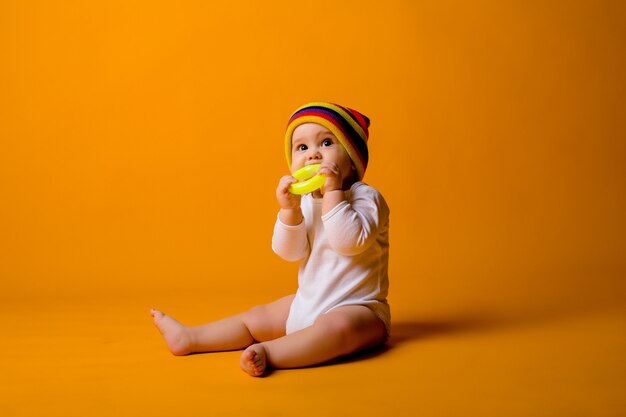 bambino in una tuta bianca e cappello multicolore in possesso di un giocattolo seduto su un muro giallo