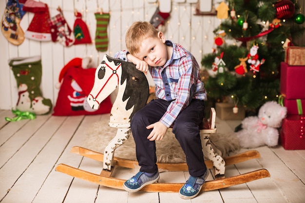 Bambino piccolo che monta cavallo a dondolo di legno davanti all'albero di Natale