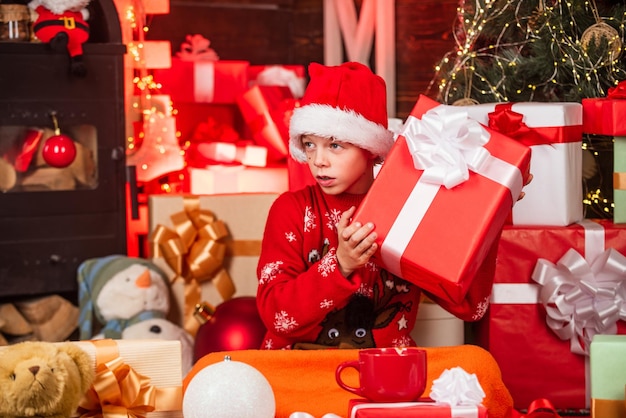 Bambino piccolo ragazzo felice trovare regali vicino all'albero di Natale Negozio di articoli da regalo Bambino cappello da Babbo Natale scatole regalo di Natale Buon Natale Bellezza dell'inverno Tempo libero e attività Santo Stefano Incuriosito carino Babbo Natale