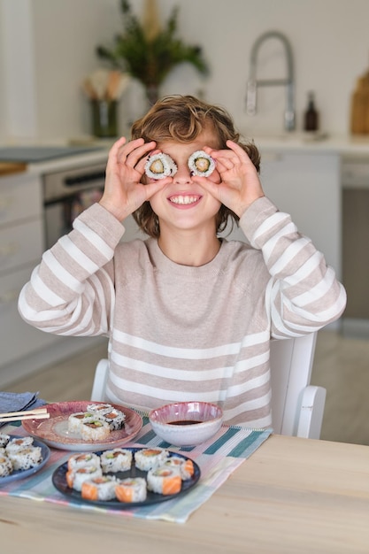 Bambino preadolescente che sorride e mette il sushi sugli occhi mentre è seduto a tavola contro il piatto con un set di rotoli