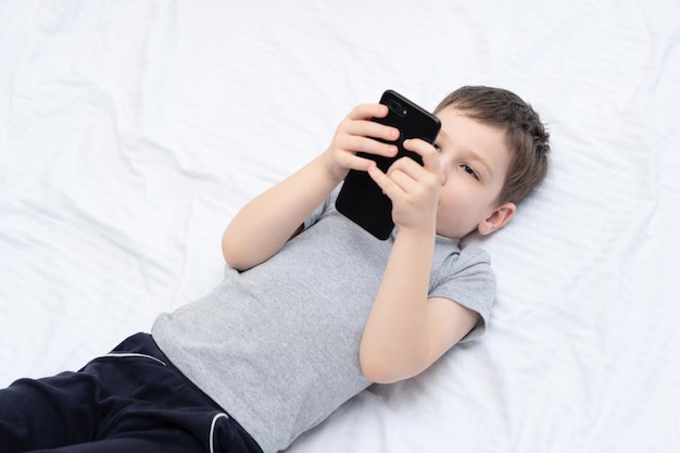 Bambino sdraiato sul letto con lo smartphone che gioca o studia qualcosa su di esso Concetto sociale e tecnologico