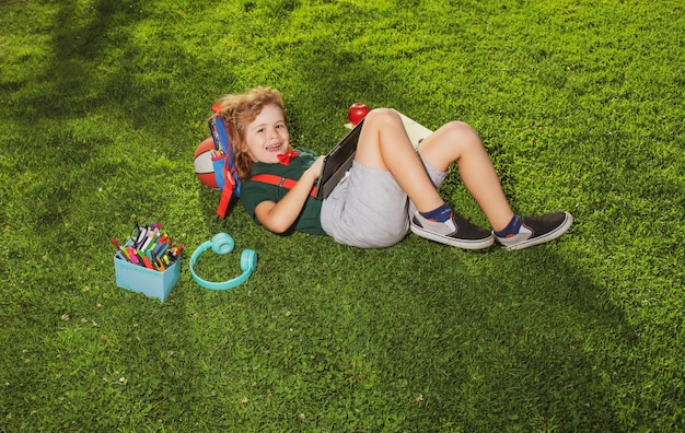 Bambino seduto sull'erba con tablet e materiale scolastico nel parco Scuola all'aperto