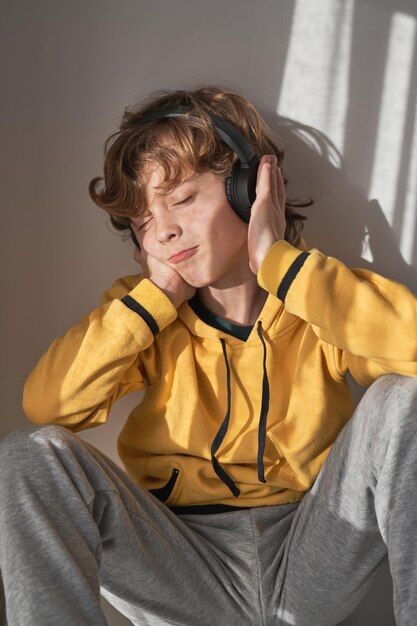 Bambino soddisfatto con le cuffie, maglione giallo e pantaloni grigi seduto con gli occhi chiusi e ascoltando musica mentre si rilassa da solo a casa