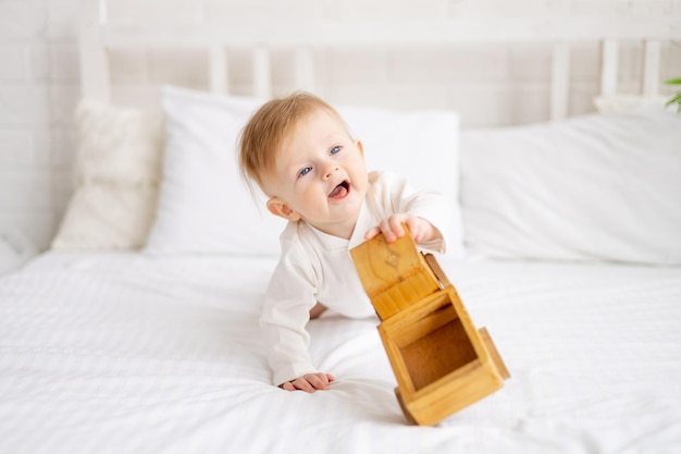 bambino sorridente 6 mesi ragazzo biondo è seduto su un grande letto in una camera da letto luminosa e gioca con una macchina giocattolo di legno in un bodysuit di cotone il concetto di beni per bambini