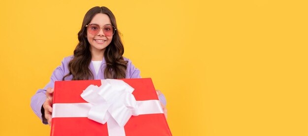 Bambino sorridente alla moda in occhiali da sole e vestito tenere la casella presente su sfondo giallo regalo Ragazza bambino con poster orizzontale regalo Intestazione banner con spazio per la copia