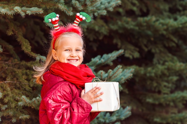 Bambino sorridente con un cerchio di Natale che tiene una scatola regalo vicino all'albero di Natale