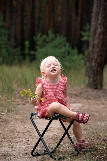 Bambino sorridente felice con wildflower. Ragazza bionda seduta su una sedia con fiore in mano nella foresta. Umore estivo.