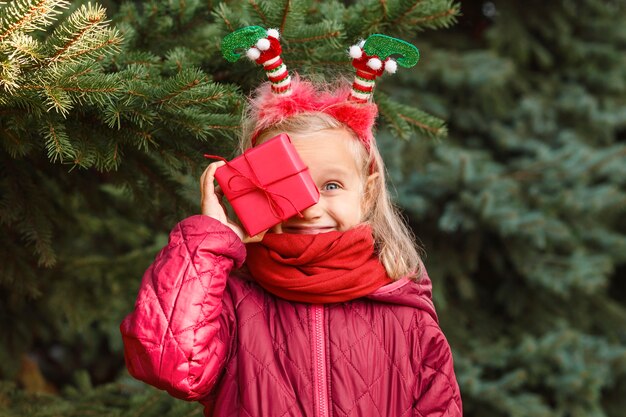 Bambino sorridente vestito di Natale che tiene una scatola regalo sullo sfondo dell'albero di Natale