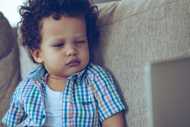 Bambino sospettoso. Piccolo bambino africano che guarda il suo laptop e sembra sospettoso mentre è seduto sul divano di casa