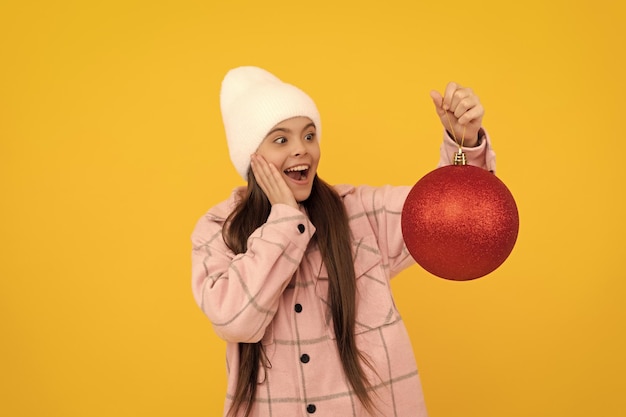 Bambino stupito in cappello invernale tenere palla decorativa di natale su sfondo giallo vacanza