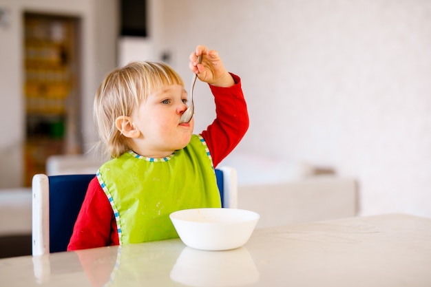 Bambino sveglio che si siede sulla sedia dei bambini alti e mangia verdura da solo nella cucina bianca.