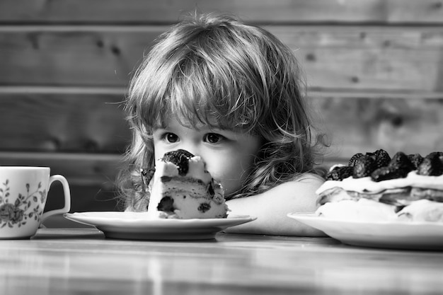 Bambino sveglio del ragazzino con lunghi capelli biondi che mangia gustosa torta cremosa o torta con frutta rossa della fragola sul tavolo con una tazza di tè