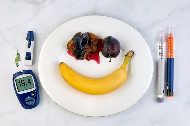 Banana e prugne tritate a forma di faccina sorridente su un piatto bianco penne per insulina e un glucometro con un livello di glucosio nel sangue molto alto